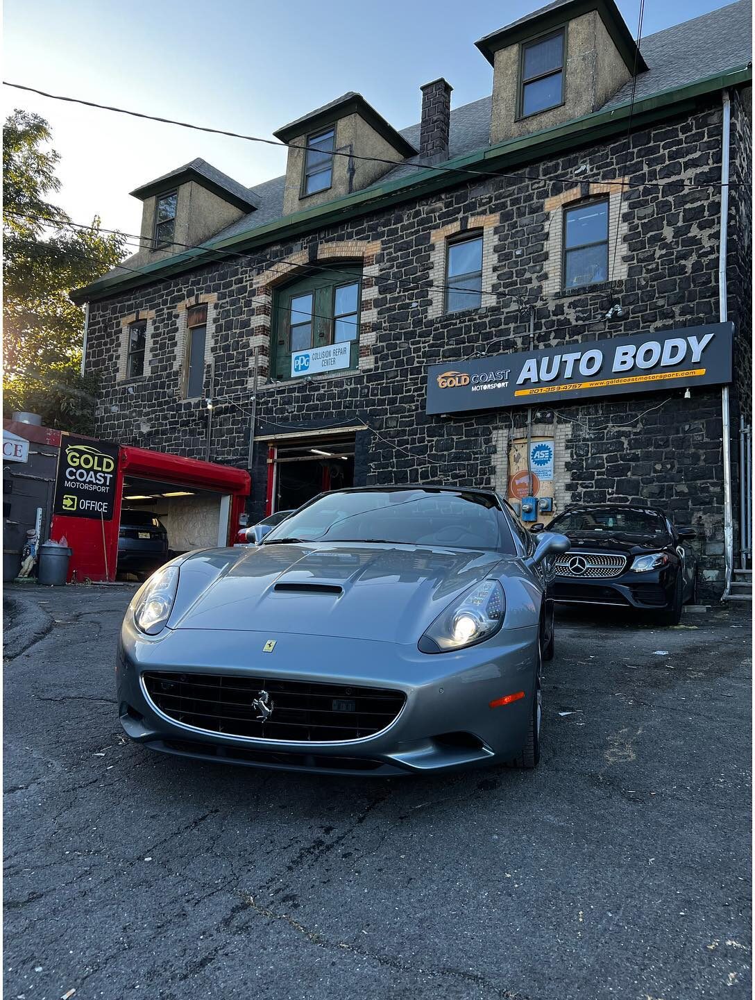 Silver-grey Ferrari California in the shop lot in front of the auto body sign.