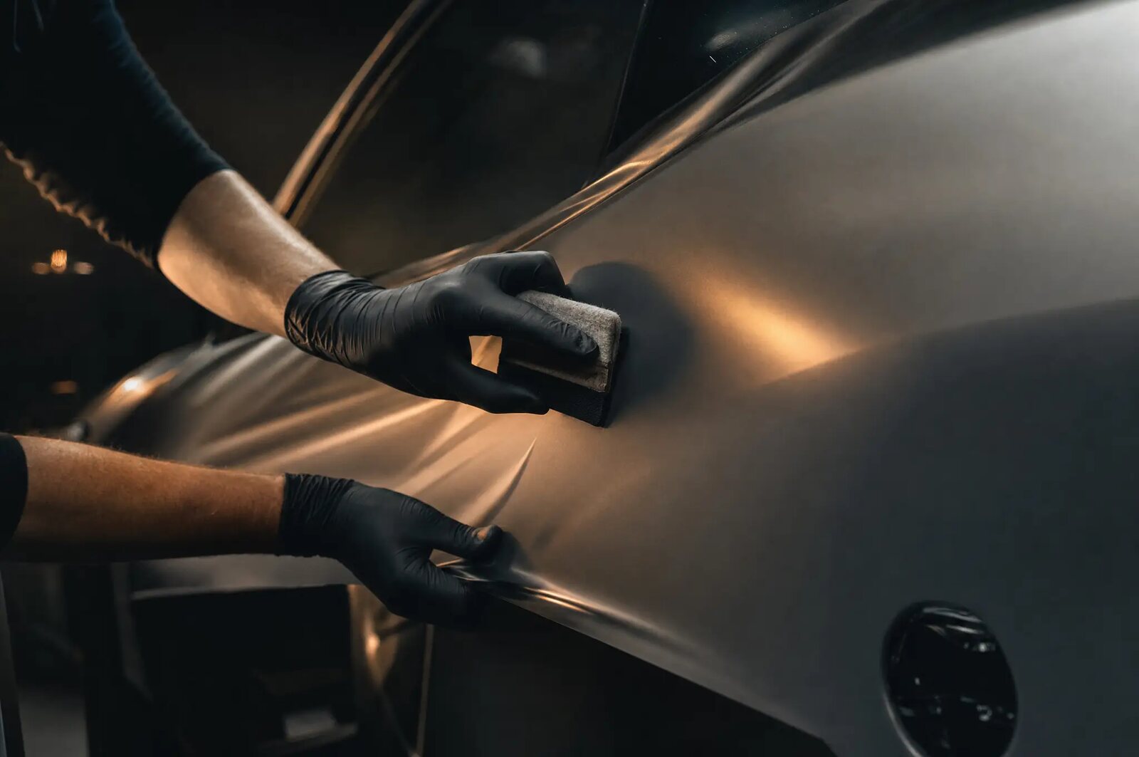 Gloved hands smoothing a satin dark graphite vinyl film onto a car door panel with a felt squeegee.
